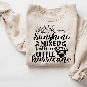 May include: A cream-colored sweatshirt with the text "Sunshine Mixed with a Little Hurricane" in black script. The design includes a sun, clouds, and a tornado graphic. The sweatshirt has long sleeves and a crew neck.