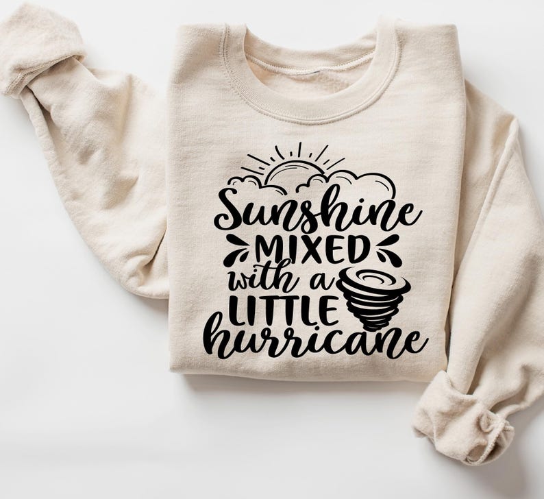 May include: A cream-colored sweatshirt with the text "Sunshine Mixed with a Little Hurricane" in black script. The design includes a sun, clouds, and a tornado graphic. The sweatshirt has long sleeves and a crew neck.