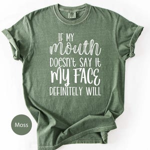 May include: Moss green t-shirt with the text "IF MY MOUTH DOESN'T SAY IT MY FACE DEFINITELY WILL" in white script. The shirt has a crew neck and short sleeves. The shirt is made of a soft, comfortable material.