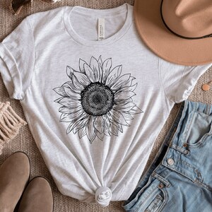 May include: A white t-shirt with a black line drawing of a sunflower on the front. The shirt is laid out on a brown surface with a pair of brown shoes, a brown hat, and a pair of blue jeans.