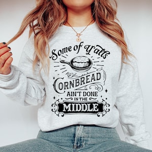 May include: A gray sweatshirt with a black graphic design that says "Some of Y'all's Cornbread Ain't Done in the Middle". The graphic features a skillet with cornbread inside.