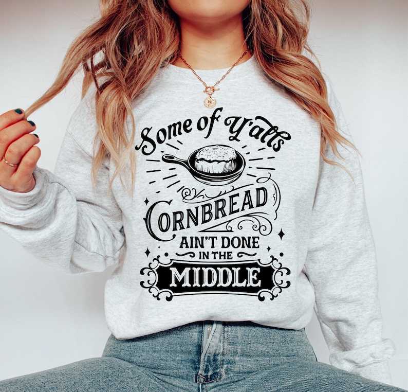 May include: A gray sweatshirt with a black graphic design that says "Some of Y'all's Cornbread Ain't Done in the Middle". The graphic features a skillet with cornbread inside.