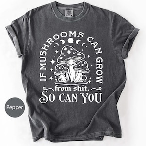 May include: A dark gray t-shirt with a white graphic of three mushrooms and the text "IF MUSHROOMS CAN GROW from shit, SO CAN YOU". The shirt is made of a soft material and has rolled-up sleeves.