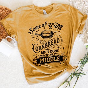May include: A mustard yellow t-shirt with a black graphic that reads "Some of Y'alls Cornbread Ain't Done in the Middle". The graphic features a vintage-style font and a black silhouette of a skillet with cornbread inside.