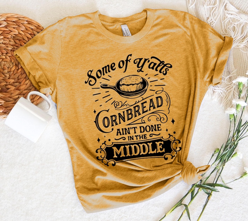 May include: A mustard yellow t-shirt with a black graphic that reads "Some of Y'alls Cornbread Ain't Done in the Middle". The graphic features a vintage-style font and a black silhouette of a skillet with cornbread inside.