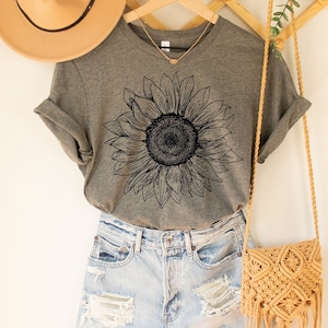 May include: A gray t-shirt with a black line drawing of a sunflower on the front. The shirt is paired with light blue denim shorts and a tan crocheted bag.