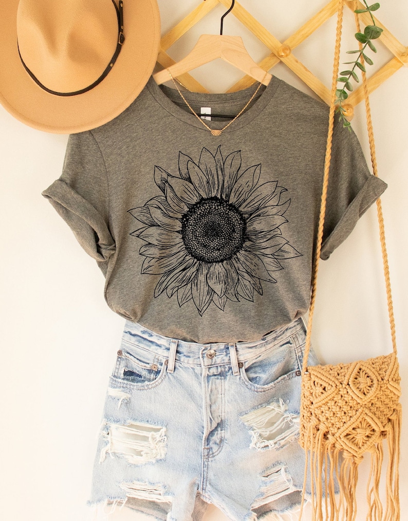 May include: A gray t-shirt with a black line drawing of a sunflower on the front. The shirt is paired with light blue denim shorts and a tan crocheted bag.