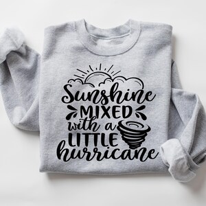 May include: A light gray sweatshirt with the text "Sunshine mixed with a little hurricane" in black script. The design includes a sun, clouds, and a tornado graphic. The sleeves are cuffed.