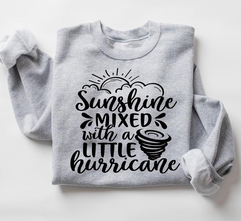 May include: A light gray sweatshirt with the text "Sunshine mixed with a little hurricane" in black script. The design includes a sun, clouds, and a tornado graphic. The sleeves are cuffed.