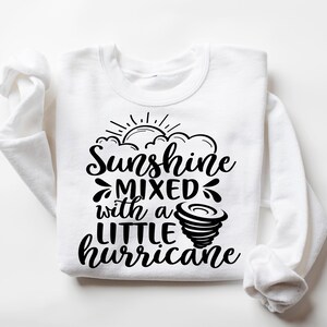 May include: White sweatshirt with the phrase "Sunshine mixed with a LITTLE hurricane" in black script. The design includes a sun, clouds, and a tornado graphic. The sweatshirt has long sleeves and a crew neck.