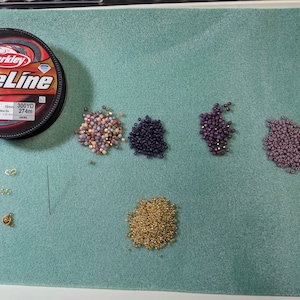 May include: A spool of Berkley FireLine fishing line, 8lb test, 300 yards, 274m, with various colours of seed beads in small piles on a green surface. The beads are gold, pink, purple, and dark purple.
