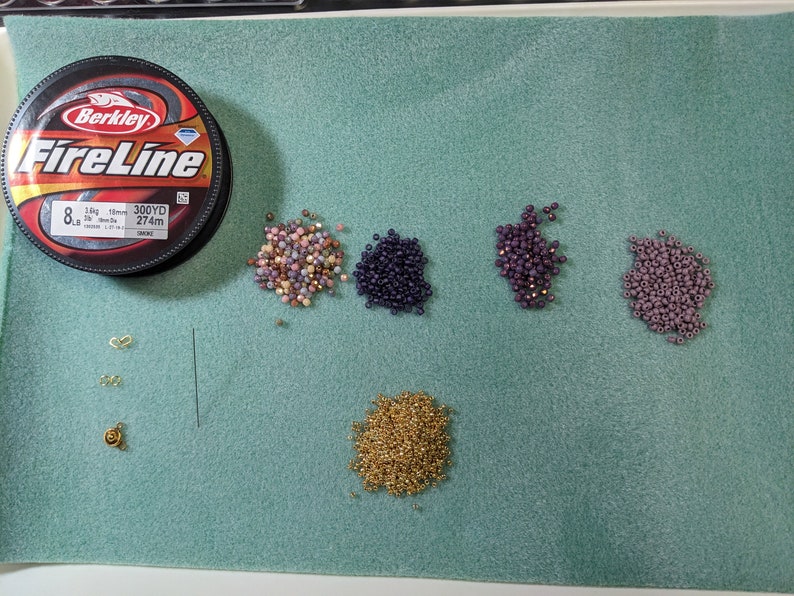 May include: A spool of Berkley FireLine fishing line, 8lb test, 300 yards, 274m, with various colours of seed beads in small piles on a green surface. The beads are gold, pink, purple, and dark purple.