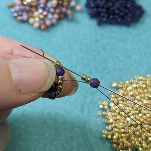 May include: Close-up of a hand holding a beaded bracelet with gold and purple beads. The bracelet is being strung with a needle and thread. There are piles of beads in the background.