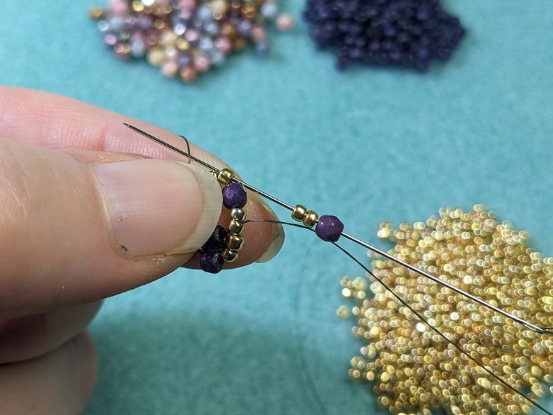May include: Close-up of a hand holding a beaded bracelet with gold and purple beads. The bracelet is being strung with a needle and thread. There are piles of beads in the background.