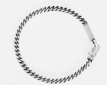 May include: A silver-colored curb chain bracelet. The bracelet features a series of interlocking, rounded links. The clasp is a rectangular bar with a hinged closure. The jewelry is displayed against a white background.