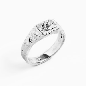 May include: A silver signet ring with a textured finish and a small bird design on the top.