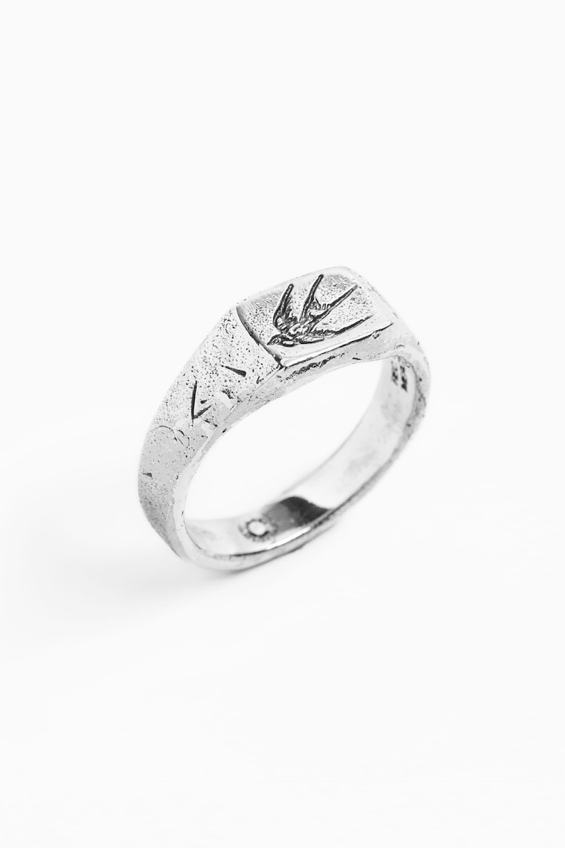May include: A silver signet ring with a textured finish and a small bird design on the top.