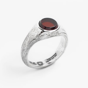 Puede incluir: Un anillo de plata con una banda texturizada y una gema grande, ovalada y de color rojo oscuro. El anillo tiene un diseño moderno y minimalista, adecuado para el uso diario. La gema está engastada en un bisel liso, que contrasta con la textura rugosa de la banda.