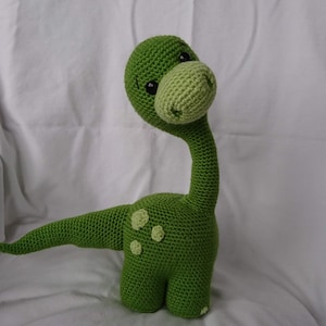 May include: A green crocheted dinosaur toy with black eyes and a long neck. The dinosaur has a light green nose and spots on its body.