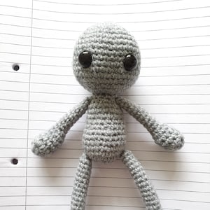 Könnte beinhalten: Eine graue gehäkelte Amigurumi-Puppe mit schwarzen Knopfaugen. Die Puppe hat ein einfaches Design mit langen Armen und Beinen.
