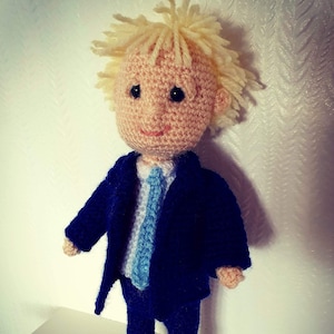 May include: A crocheted doll with blonde hair wearing a blue suit and a light blue tie.