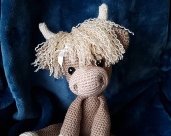 May include: A crocheted stuffed toy cow with brown fur, white hooves, and a white bow. The cow has a fluffy, light brown mane.