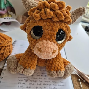 May include: A handmade, plush Highland cow amigurumi. The crochet cow features a brown body, tan horns and hooves, and large, reflective black eyes. The cow is sitting on a white surface with a notebook and pen nearby. A ball of orange yarn is in the background.