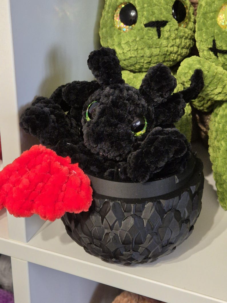 May include: A black crocheted dragon plush toy sitting in a black egg-shaped container with a red crocheted wing. The dragon has large black eyes and a green mouth.