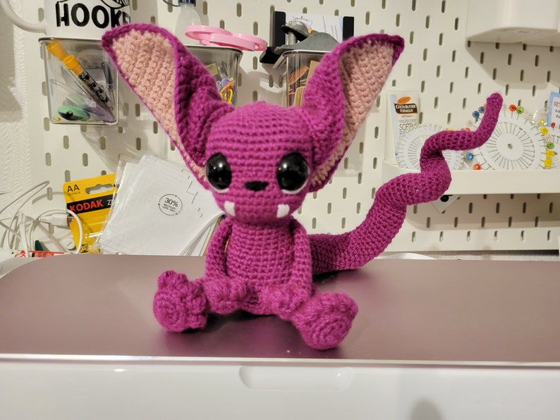 Puede incluir: Una criatura de crochet de color morado y rosa con ojos grandes y una cola larga. La criatura est&aacute; sentada sobre una superficie blanca.