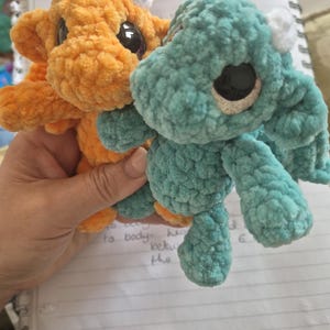 May include: Two handmade crochet creatures, one orange and one teal. They have large black eyes and small white horns. The creatures are held in a hand, resting on a notebook with handwritten notes and a red crochet hook.