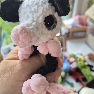 May include: A handmade crochet cow plushie with a white body, black ears, and a pink snout and feet. The cow has large, sparkly eyes and a curly tail. The plushie is held in a hand.
