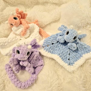 May include: A collection of handmade crochet dragon security blankets in pink, purple, and blue. Each dragon has large eyes and a soft, plush texture. The blankets have a white border and are set on a fluffy white surface.