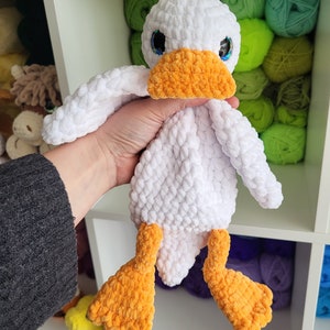 May include: White crocheted duck plush toy with orange beak and feet. The duck has blue eyes and is sitting on a shelf with colorful yarn balls.