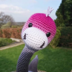 May include: A crocheted stuffed animal shaped like a long-necked dinosaur. The dinosaur is pink and white with black eyes and a black nose. The dinosaur's neck is a gradient of gray and black.