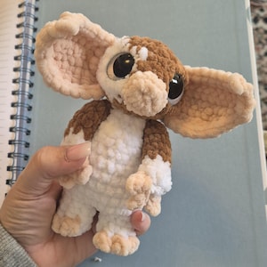 May include: A plush crochet creature featuring large, rounded ears, a white belly, and brown patches on the head and body. The figure has oversized, glossy black eyes and a textured, soft yarn construction with small limbs and feet.