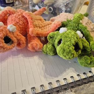 May include: Crocheted dragon toys in shades of orange and green, with white accents and large black eyes. The plush toys are lying on a spiral notebook, showcasing their soft, textured appearance. A handmade craft item.