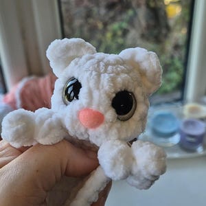 May include: A handmade white crochet bunny with large black eyes and a pink nose. The plush toy has small ears and outstretched arms, giving it a friendly appearance. The bunny is made of soft, textured yarn.
