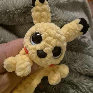 May include: A small, handmade crochet plush toy made of soft, pale yellow yarn. It features two long ears with black tips, large round black plastic eyes, a small black nose, and two small red circular patches on its cheeks. The texture is fuzzy.