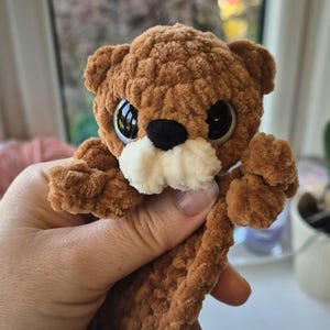 May include: A handmade brown plush otter with large, expressive eyes and a white muzzle. The plush toy is held in a hand, showcasing its soft, textured material. The otter's design includes small arms and a rounded body, perfect for a child's toy or nursery decor.