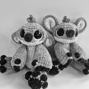 Può includere: Due peluche fatti a mano all'uncinetto, simili a un personaggio dei cartoni animati, sono esposti. I giocattoli sono grigi con accenti neri su naso, occhi e piedi. Hanno grandi occhi rotondi e orecchie flosce e sono seduti uno accanto all'altro.