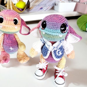May include: Two crocheted amigurumi toys. The first is a rainbow coloured creature with large eyes and a pink and purple body. The second is a purple and blue creature with a white vest and red trainers.