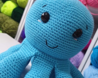 May include: A crocheted blue octopus with black eyes and a stitched smile. The octopus is sitting on a white surface.