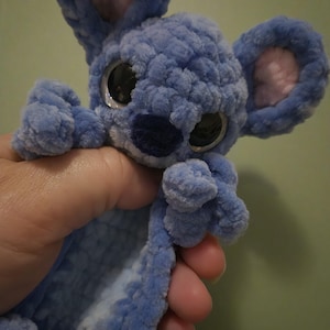 May include: A small, blue, crocheted plush toy with a soft, fuzzy texture. It features large, round, black eyes, a dark blue nose, and two prominent, upright ears with light pink centres. The character has a rounded head and small limbs.