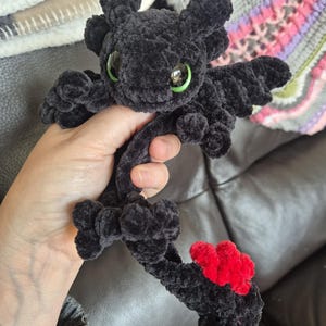 May include: A black plush dragon toy made of soft, fuzzy yarn with large, bright green eyes. The creature features small wings, textured ears, and a long tail ending in a vibrant red, heart-shaped fin detail. The construction is entirely crocheted.