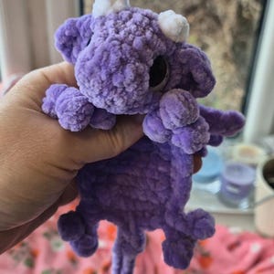 May include: A handmade, plush purple dragon toy with white horns and large black eyes. The dragon has small wings and a curled tail. The toy is made of a soft, textured material, and is held in a person's hand.