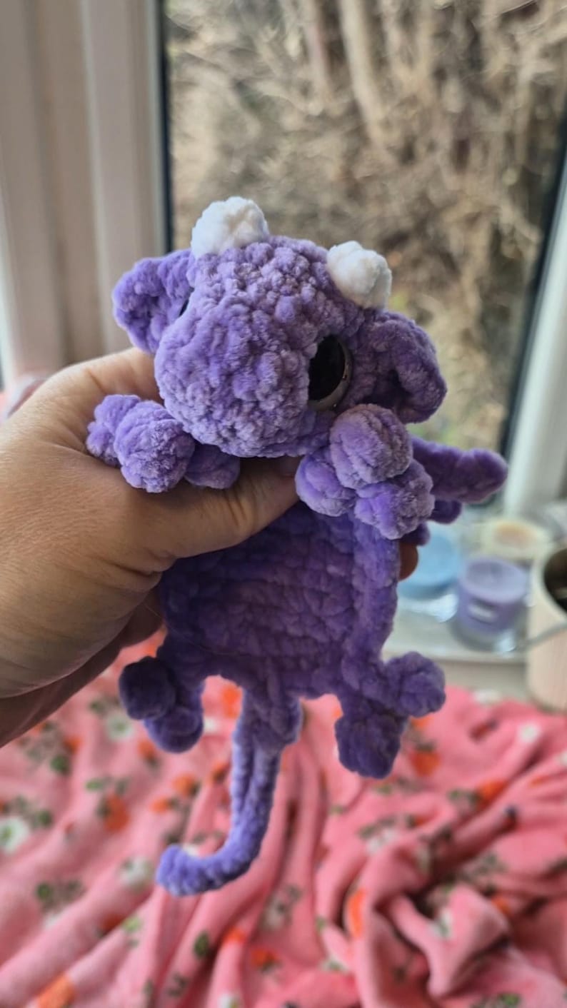 May include: A handmade, plush purple dragon toy with white horns and large black eyes. The dragon has small wings and a curled tail. The toy is made of a soft, textured material, and is held in a person's hand.