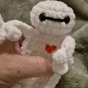 May include: A white crocheted plush toy featuring a rounded head with two black oval eyes connected by a thin black line. The body is made of soft white yarn and includes a small, bright red heart stitched onto the center of the chest area.