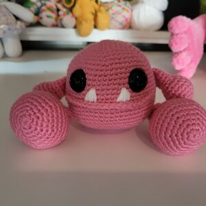 May include: A pink crocheted monster with black eyes and white teeth. The monster has two round pink arms.