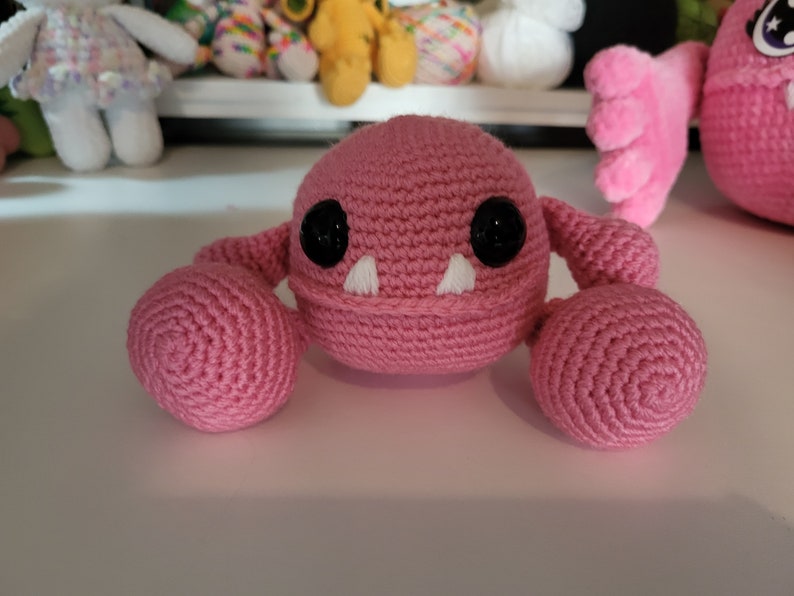 May include: A pink crocheted monster with black eyes and white teeth. The monster has two round pink arms.