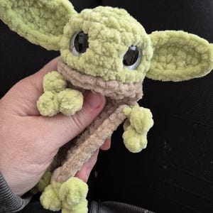 May include: A plush toy crafted from light green and tan yarn, featuring large, dark, glossy eyes and prominent, wide ears. The creature has a textured, soft appearance with small, rounded limbs and a distinct collar-like section in a tan shade.
